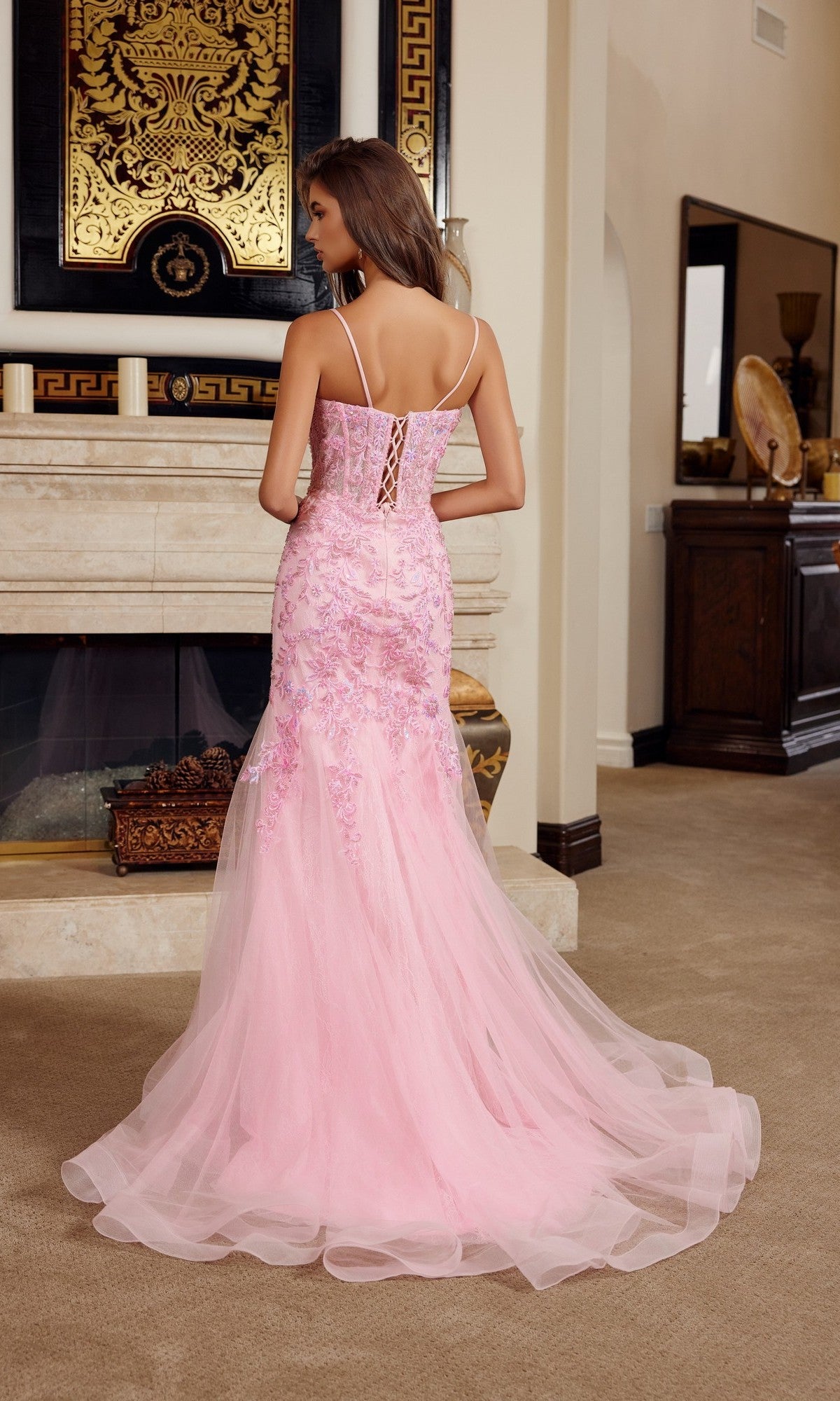 Wearing the Nox Anabel H1874, a long pink lace and tulle prom dress with beaded sequin embroidery, a woman stands with her back to the camera in a tastefully decorated living room.